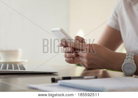 Close up image of modern cellphone in womans hand at desk with laptop. Office worker boring at workplace and playing games, writing in social networks. Businesswoman looking important contact on phone