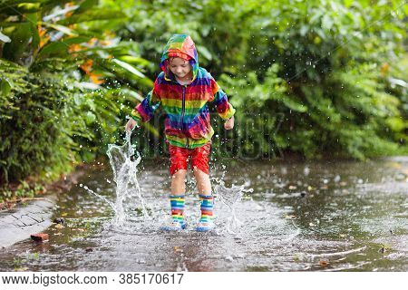 Kid Playing In The Rain In Autumn Park. Child Jumping In Muddy Puddle On Rainy Fall Day.