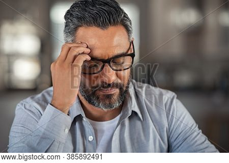 Frustrated middle aged man sitting on couch at home. Close up face of stressed indian businessman wearing eyeglasses with eyes closed. Overworked middle eastern business man with terrible migraine.