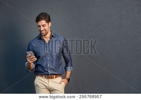 Portrait of young man leaning against a grey wall using mobile phone. Happy business man messaging with smartphone isolated on gray background with copy space. Smiling guy typing and reading a message