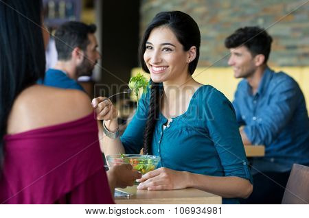 Closeup shot of young women eating salad at restaurant. Happy female friends smiling and chatting. Portrait of smiling girl holding a forkful of salad during lunch break.