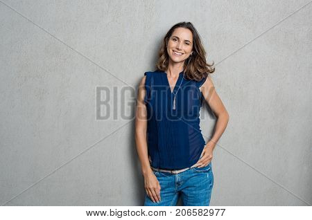 Portrait of cheerful mature woman standing against grey wall. Happy mid woman looking at camera against grey background with copy space. Smiling carefree latin woman looking at camera.