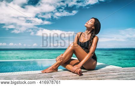 Swimsuit model beauty skincare suntan Asian woman posing sunbathing tanning in black one piece bathing suit. Beautiful girl relaxing at overwater infinity pool luxury resort. Spa, wellness, laser.