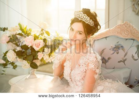 Bride. Wedding. The bride in a short dress with lace in the crown earrings in luxurious interior. Wedding bouquet, makeup, hairstyle. Wedding Style