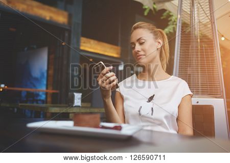 Charming European female browsing wifi via mobile phone during morning breakfast in cafe bar beautiful woman reading text message on her cell telephone while sitting in coffee shop during free time