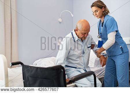 Lovely nurse assisting senior man to get up from bed at hospital. Caring nurse supporting elderly patient while getting up from bed and move towards wheelchair at nursing home. 