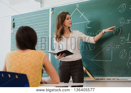 Teacher or docent or educator giving while lesson in front of a blackboard or board a sheet of paper and educate or teaching students or pupils or mates in a school or class.