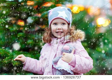 Cute little smiling kid girl with christmas tree. Happy child in winter clothes and toy choosing xmas tree on Christmas market with lights on background. Family, tradition, celebration concept