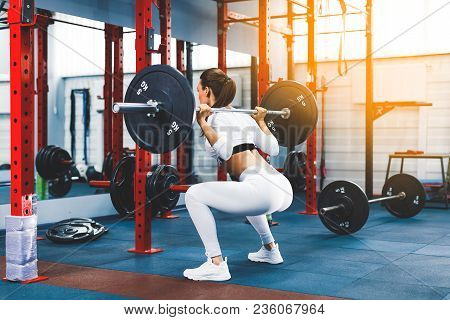 Back View Young Adult Girl Doing Squat In Gym With Barbell. Fit Woman In Great Shape. The Developmen