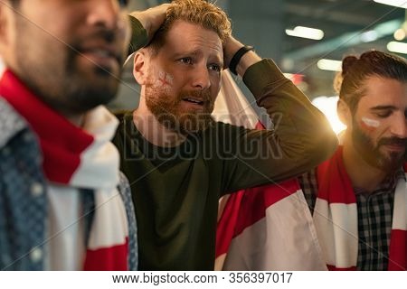 Soccer fans feeling disappointed on seeing favourite team lose the match. Group of disappointed supporter wearing scarf and paint on face upset. Football fans with teamâs flag upset about losing game.