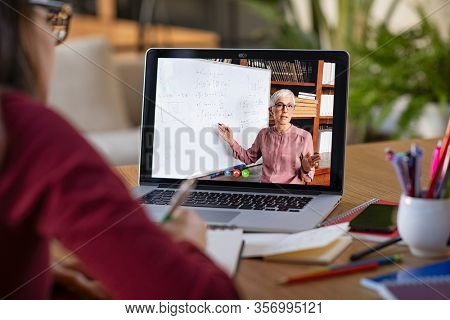 Young student watching lesson online and studying from home. Young woman taking notes while looking at computer screen following professor doing math on video call. Girl studying from home on pc.