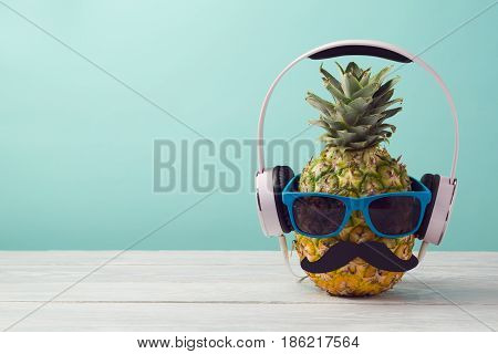 Pineapple with sunglasses and headphones on wooden table over mint background. Tropical summer vacation and beach party concept.