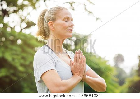 Mature woman in park joining hands with closed eyes and meditates. Relaxed senior woman with joined hands breath deeply with closed eyes. Healthy woman meditating outdoor.