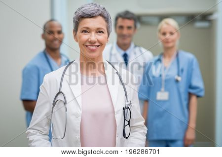 Portrait of smiling woman doctor standing in hospital with team in background. Senior female doctor in front of her medical staff. Team of medical professionals looking at camera.