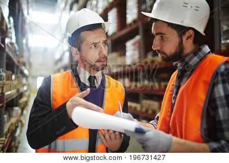 Portrait of warehouse worker talking to supervising manager while doing inventory control in entrepotTwo Warehouse Workers Reviewing Goods