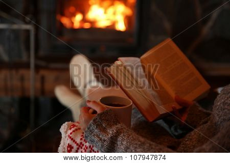 Woman resting with cup of hot drink and book near fireplace