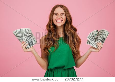 Portrait of a satisfied happy woman winner with long hair holding bunch of money banknotes in each hand isolated over pink background