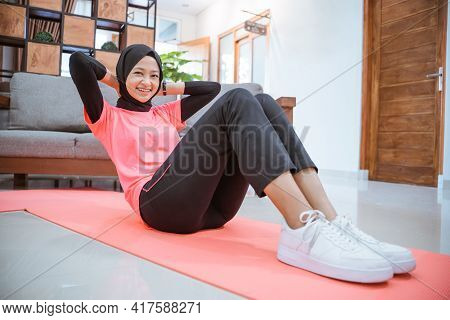 A Girl In A Hijab Gym Outfit Smiles While Doing A Sit-up On A Mat On The Floor