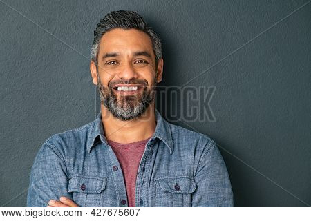 Close up face of handsome mature man with beard looking at camera. Happy mid adult indian man isolated against grey wall. Portrait of smiling mixed race guy on gray background with copy space.