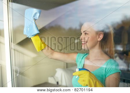 people, housework and housekeeping concept - happy woman in gloves cleaning window with rag and cleanser spray at home