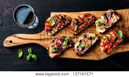 Brushetta set for wine. Variety of small sandwiches with prosciutto, tomatoes, parmesan cheese, fresh basil and balsamic creme served with glass of red wine on rustic wooden board over dark background, top view