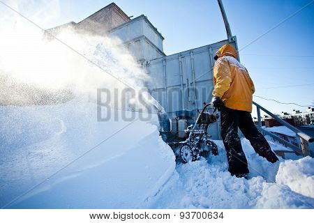 Man in yellow winter jacket cleaning garage path near his house with snow throwing machine