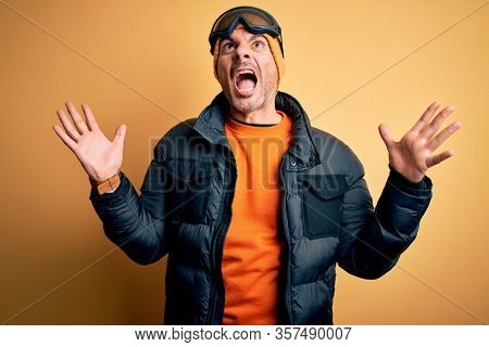 Young handsome skier man skiing wearing snow sportswear using ski goggles crazy and mad shouting and yelling with aggressive expression and arms raised. Frustration concept.