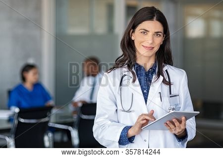 Portrait of beautiful mature woman doctor holding digital tablet and looking at camera. Confident female doctor using digital tablet with colleague talking in background at hospital. Latin nurse.