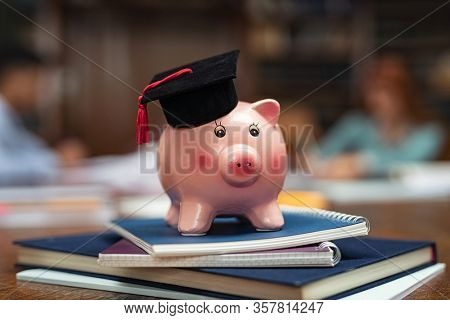 Piggy bank wearing mortar board on pile of books in library while students studying in background. Saving money for college with piggy bank on books in library. Savings and education debt concept.