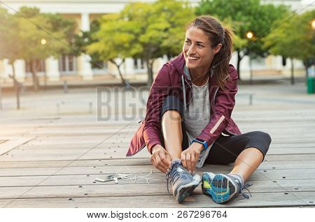 Mature fitness woman tie shoelaces on road. Cheerful runner sitting on floor on city streets with mobile and earphones wearing sport shoes. Active latin woman tying shoe lace before running.