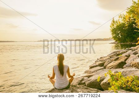 Yoga class outside in nature park by lake river shore. Woman sitting in lotus pose meditating by the water in morning sun flare sunrise. Meditation wellness.