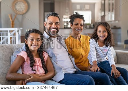 Portrait of cheerful ethnic family at home sitting on sofa and looking at camera. Happy indian family with two children relaxing at home. Mixed race parents with their daughters in new home.