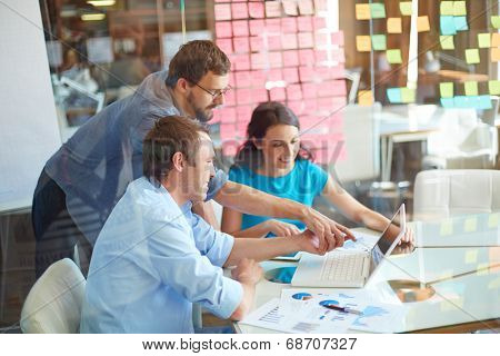 Group of three successful business partners in casual looking at laptop screen while two young men pointing at it at meeting in office