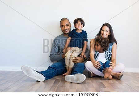 Happy multiethnic family sitting on floor with children. Smiling couple sitting with two sons and looking at camera. Mother and black father with their children leaning on wall with copy space.