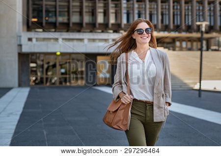 Portrait of successful happy woman on her way to work on street. Confident business woman wearing blazer carrying side bag walking with a smile. Smiling woman wearing sunglasses and walking on street.