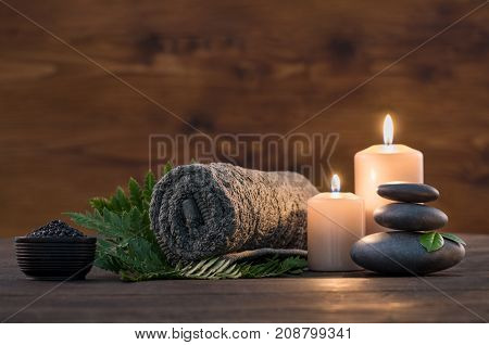 Towel on fern with candles and black hot stone on wooden background. Hot stone massage setting lit by candles. Massage therapy for one person with candle light. Beauty spa treatment and relax concept.