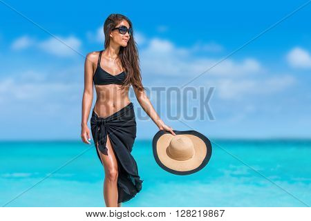 Elegant beach woman in bikini and fashion sarong standing on shore. Sexy lady in black beachwear, floppy hat, sunglasses enjoying sun on tropical destination during summer vacation in the Caribbean.