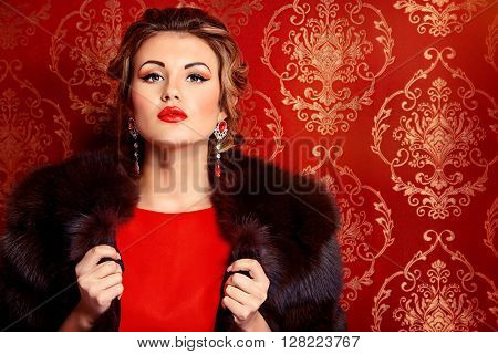 Portrait of a beautiful woman in red dress and luxurious fur coat alluring by vintage wallpaper. Luxury, rich lifestyle. Jewellery. Fashion shot. 