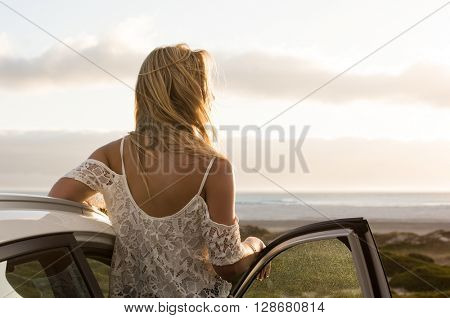 Young traveller woman enjoying sunset standing outside car. Serene woman leaning on car during sunset. Young woman on summer travel to the coast.
