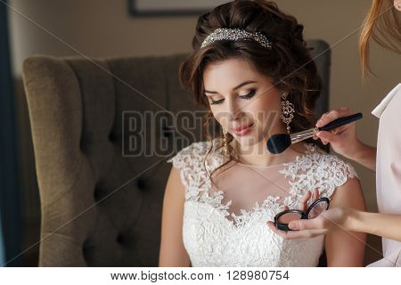 Beautiful young bride with curly brunette hair,beautiful wedding hairstyle with tiara,large earrings,white wedding dress,a gentle smile,sitting on a high chair in the hotel room, the make-up, using the services of a makeup artist