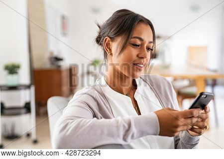 Mature african woman sitting on couch at home using smartphone. Black woman relaxing on the sofa while messaging with a cellphone at home. Happy mature indian woman typing on mobile phone.