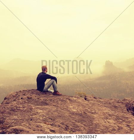 Tourist sit on peak of sandstone rock and watching into colorful mist and fog in morning valley. Sad man. Man sit. Man in jeans.Fall mountain mist. Mist in valleys. Man hand.Sad tourist sit in mist.