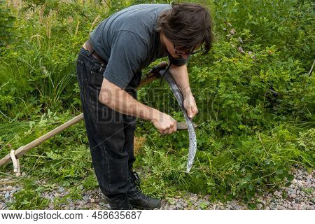 A Man Mows The Grass With A Hand Scythe. Mow The Grass With An Old Scythe. The Farmer Traditionally 