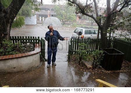 A business man stands outside in the pouring rain wearing a blue rain coat and a clear umbrella waiting for someone or the rain to stop. 