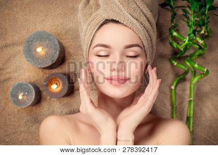 Beautiful spa woman with a towel on her head lying and touching face skin. Skincare. Beauty smiling model girl in spa salon. Skin care, acne treatment, cleansing fresh skin. Renewal cosmetics wellness