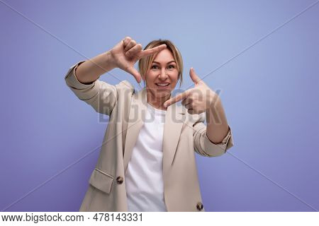 Cheerful Woman In A Jacket With Blond Hair Makes A Photo With The Help Of Her Hands On A Studio Back