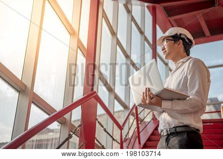 Young Asian Engineer or Architect working with laptop computer while wearing a personal protective equipment safety helmet at construction site. Engineering and building construction concepts