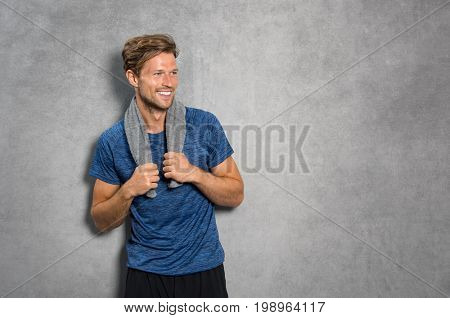 Portrait of a smiling fitness man with towel on shoulders looking away. Happy young man relaxing after training and leaning on grey wall. Sporty active guy resting after workout with copy space.