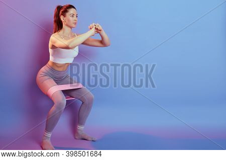 Photo Of Fitness Girl In Stylish Sportswear Squats With Rubber Band On Background Of Blue And Rose W