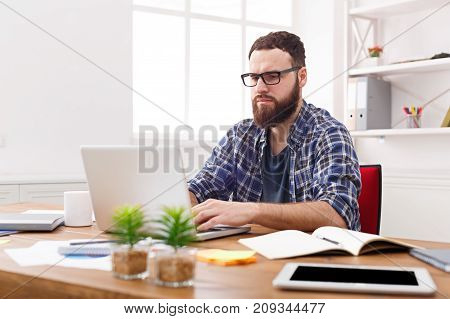 Pensive and thotful young businessman in casual wearing eyeglasses working with laptop in modern white office interior.